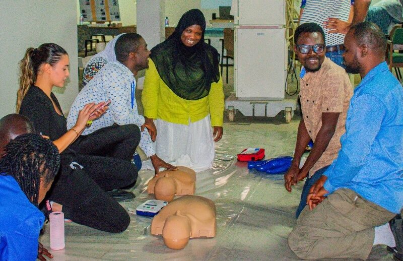 A woman wearing medical scrubs claps as four other people look at each other and smile. They are kneeling on the floor in front of human shaped dummies used for CPR training.