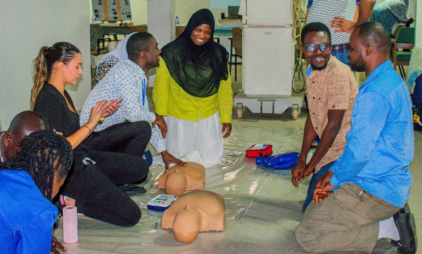 A woman wearing medical scrubs claps as four other people look at each other and smile. They are kneeling on the floor in front of human shaped dummies used for CPR training.