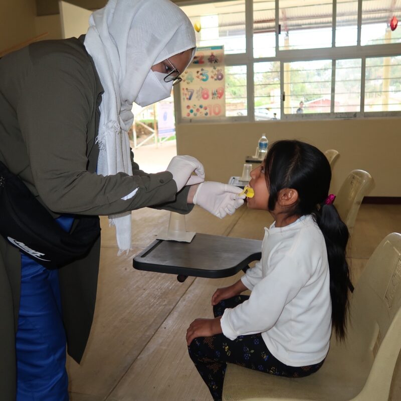 A woman wearing gloves holds a small bright yellow tray to a young girls mouth as she sits to receive this fluoride treatment.