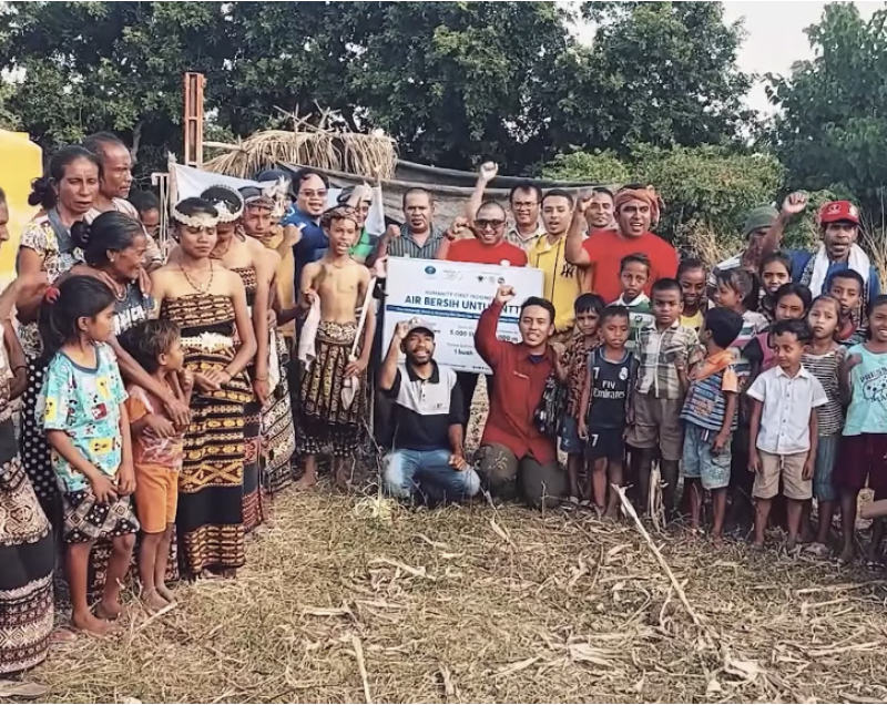 Several dozen people including some women wearing traditional Indonesian skirts pose in a group in front of their new water well in clearing of hay with trees behind them.