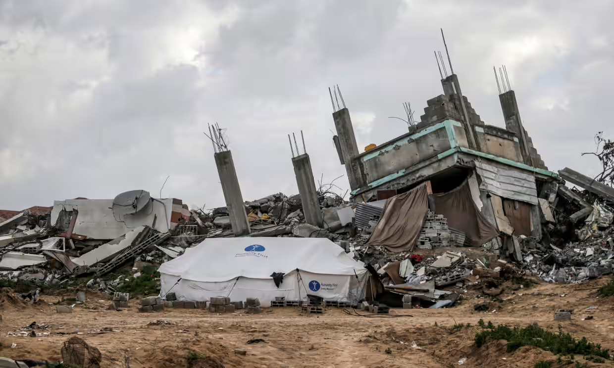 Next to a set of completely destroyed buildings on a dirt road stands a large white tent bearing the logo for Humanity First.