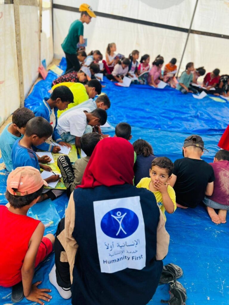 children sit in a circle on a blue tarp and write together on worksheets. Two children sit with a woman wearing a vest with the humanity first logo