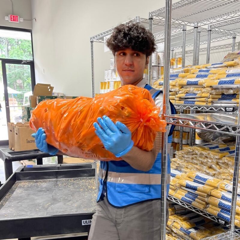 A young man stands amongst shelves of dry food goods and holds a large sack of potatos.