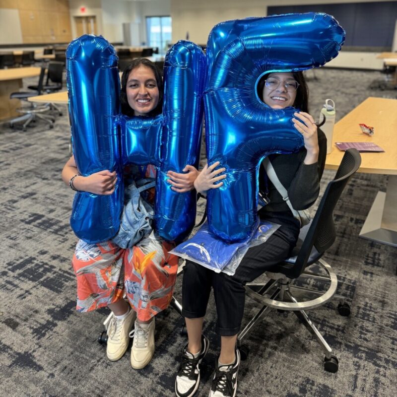 Two college aged women sit n a university classroom holding giant blue balloons. One balloon is shared as the letter H and the other is the letter F.