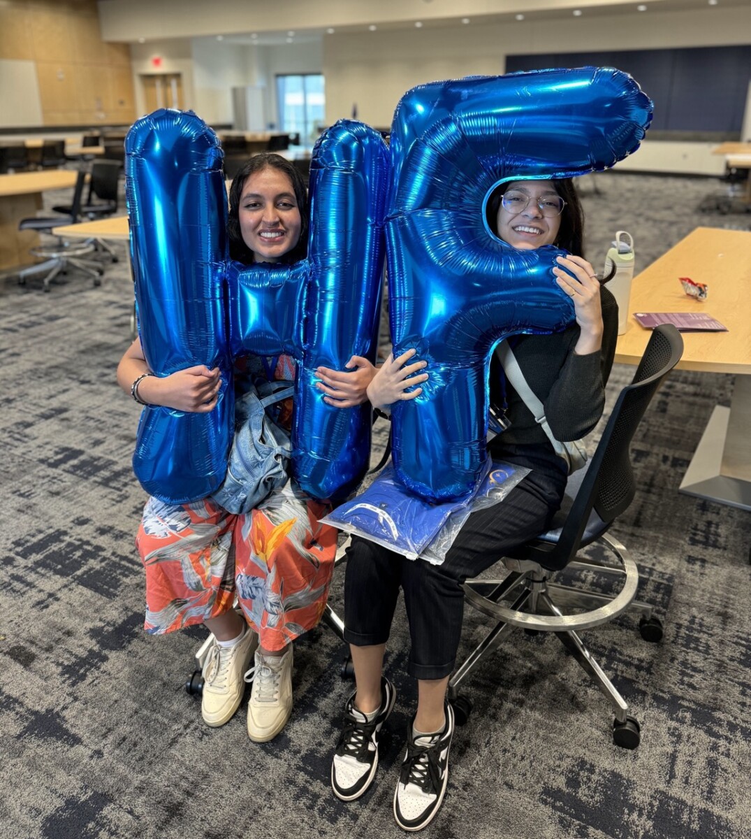 Two college aged women sit n a university classroom holding giant blue balloons. One balloon is shared as the letter H and the other is the letter F.