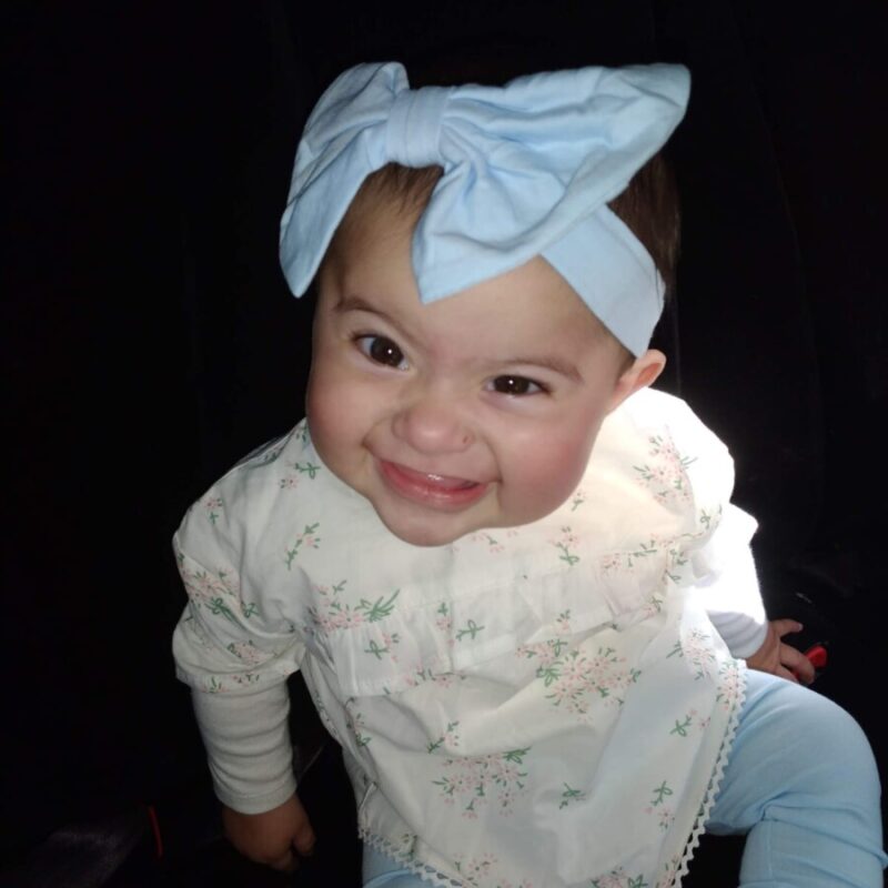 A young baby smiles and wears a blue headband with a large blue blow on her head.