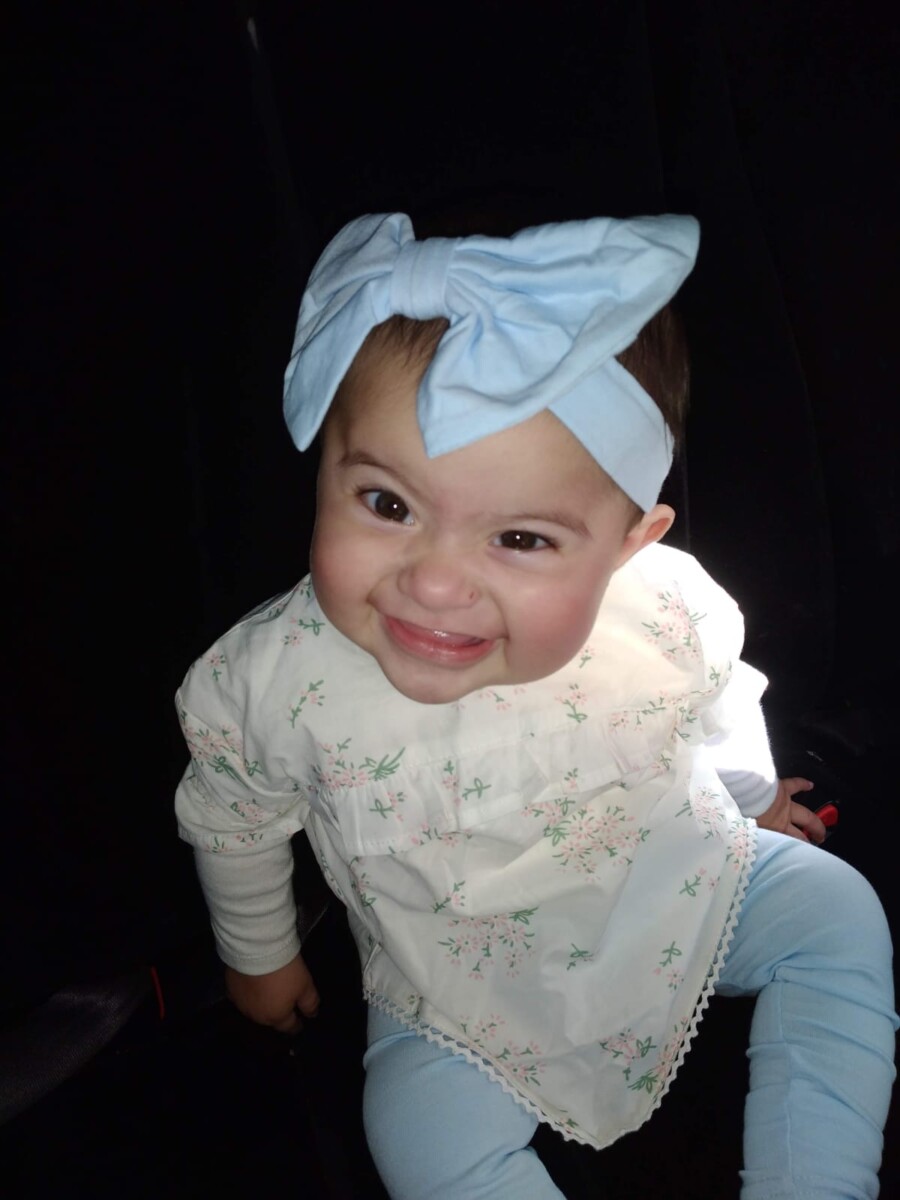 A young baby smiles and wears a blue headband with a large blue blow on her head.