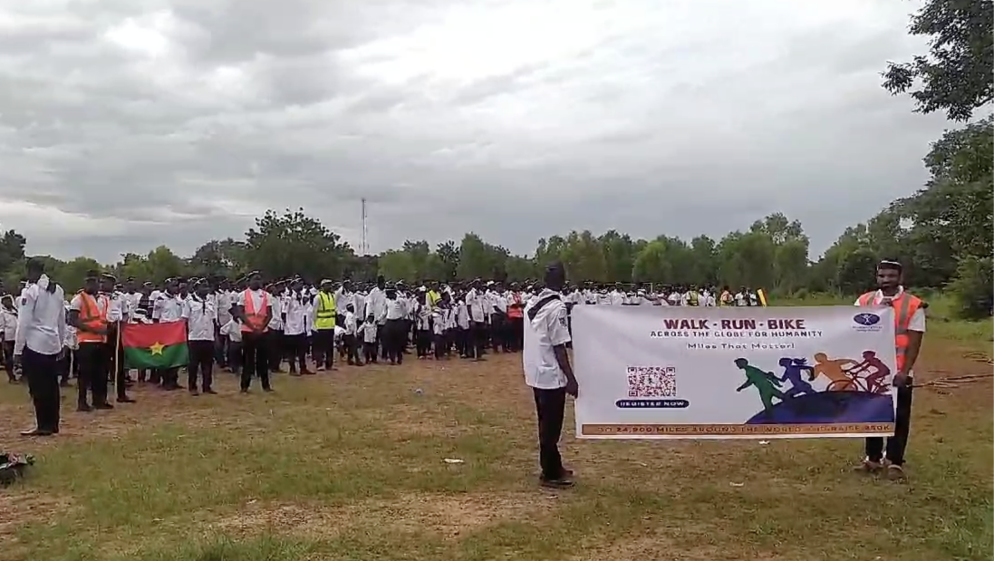 Dozens of people wearing white shirts gather in a large field. Two hold a Burkina Faso flag and two hold a banner saying Walk Run Bike