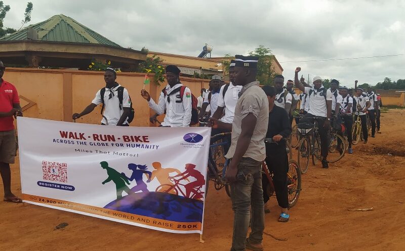 About two dozen people wear matching white shirts and wait on their bicycles. Some have their hands in the air cheering. Two people standing in the front hold a sign that says Walk Run Bike.