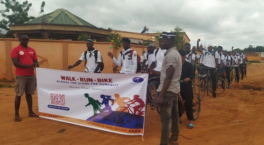 About two dozen people wear matching white shirts and wait on their bicycles. Some have their hands in the air cheering. Two people standing in the front hold a sign that says Walk Run Bike.