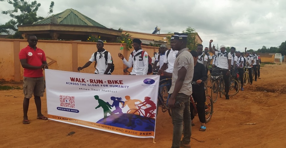 About two dozen people wear matching white shirts and wait on their bicycles. Some have their hands in the air cheering. Two people standing in the front hold a sign that says Walk Run Bike.