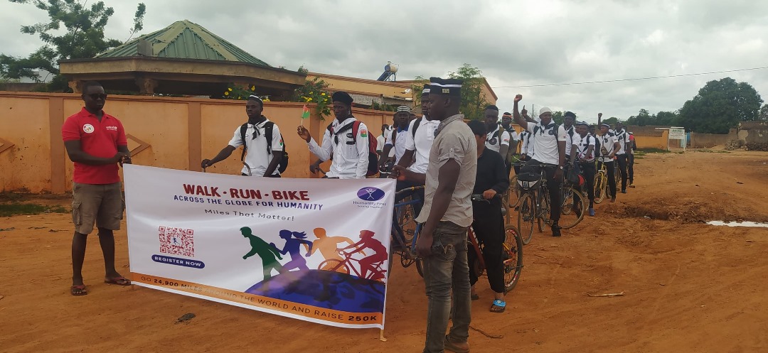 About two dozen people wear matching white shirts and wait on their bicycles. Some have their hands in the air cheering. Two people standing in the front hold a sign that says Walk Run Bike.