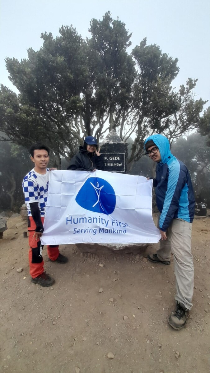 Two people hold up a banner with the Humanity First logo. They are outside with a short tree right behind them.