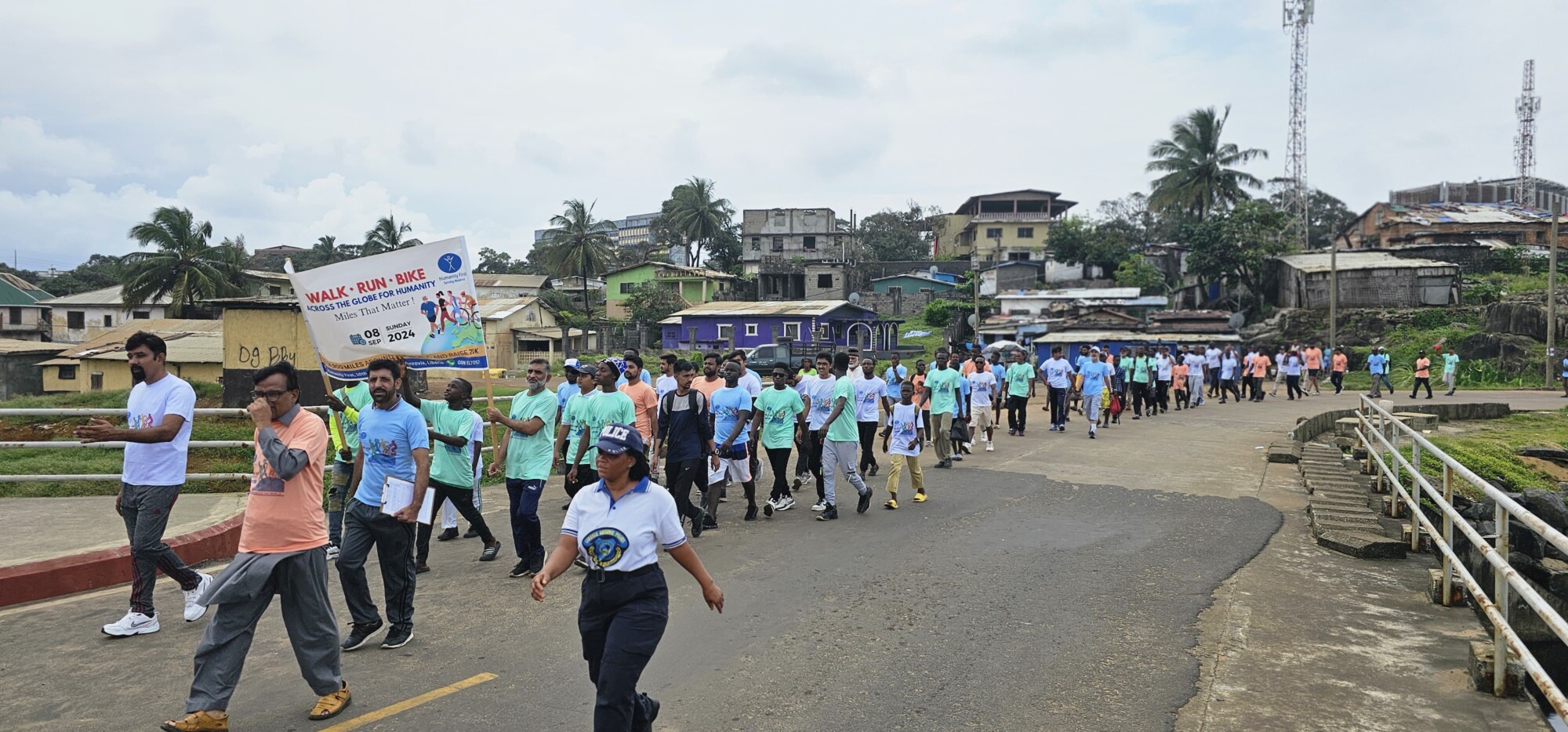 A large group of people in a long line wearing multiple different pastel colored t shirts walk outside only a road with houses behind them.