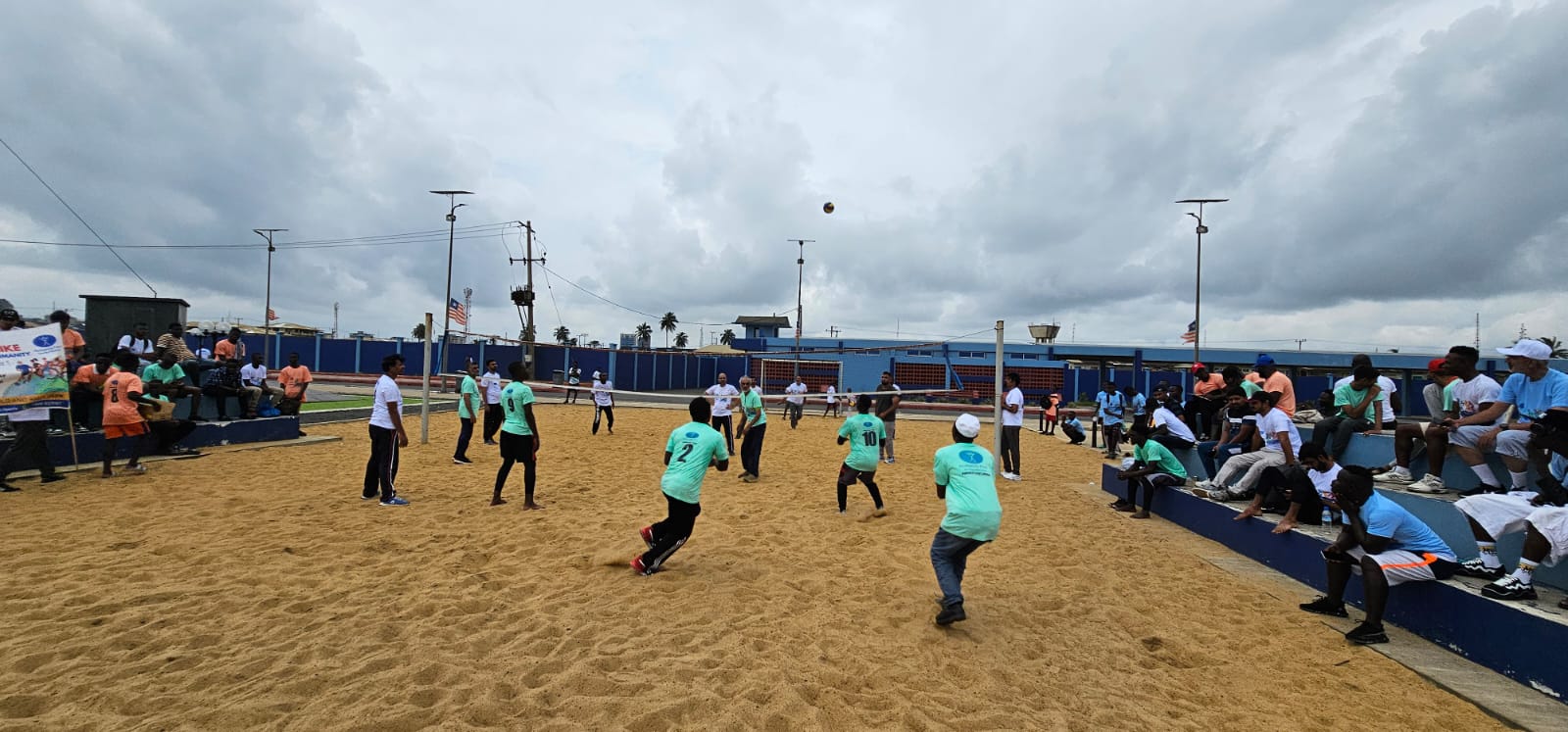 people play beach volleyball while others watch from seats on the side. A team wearing green t shirts is closest to the camera.