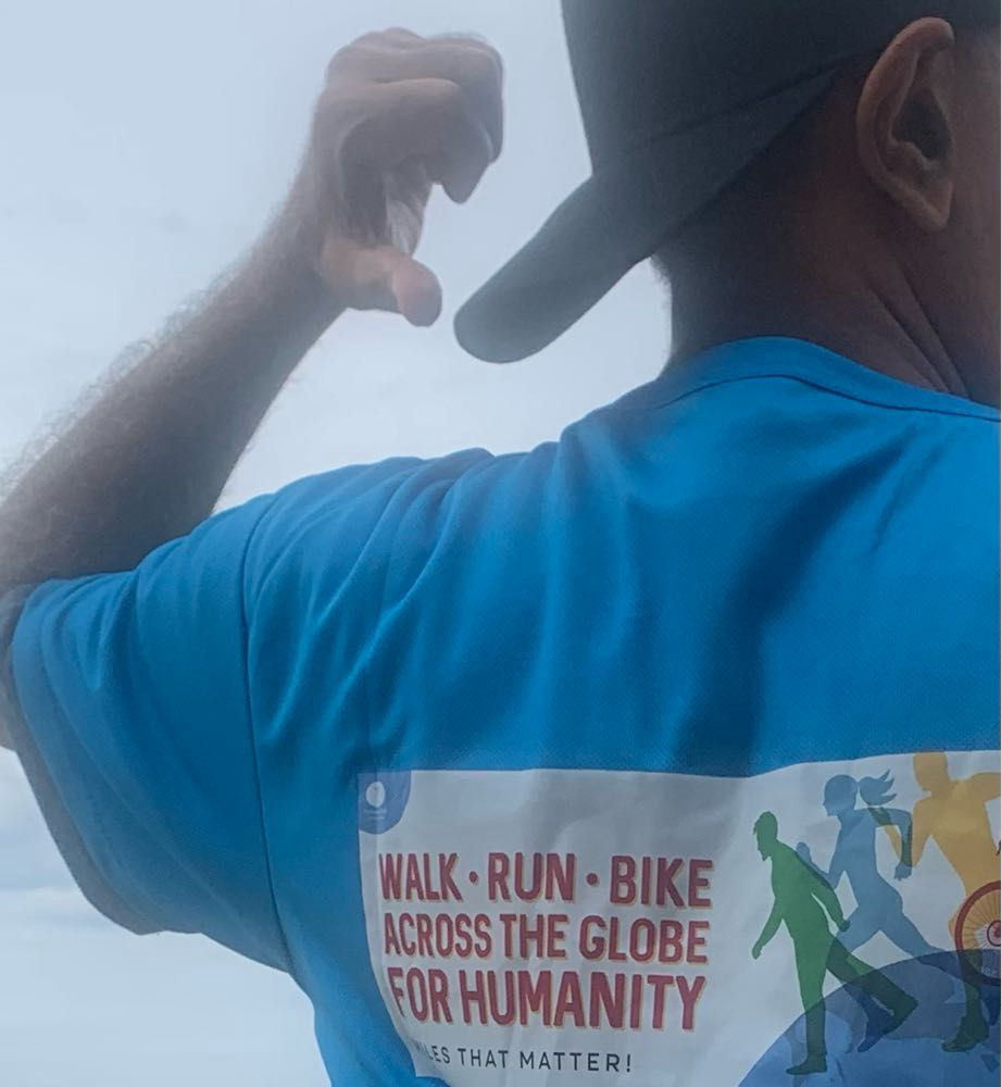 Man wearing bright blue t shirt points to the back of his shirt which says walk run bike across the globe for humanity