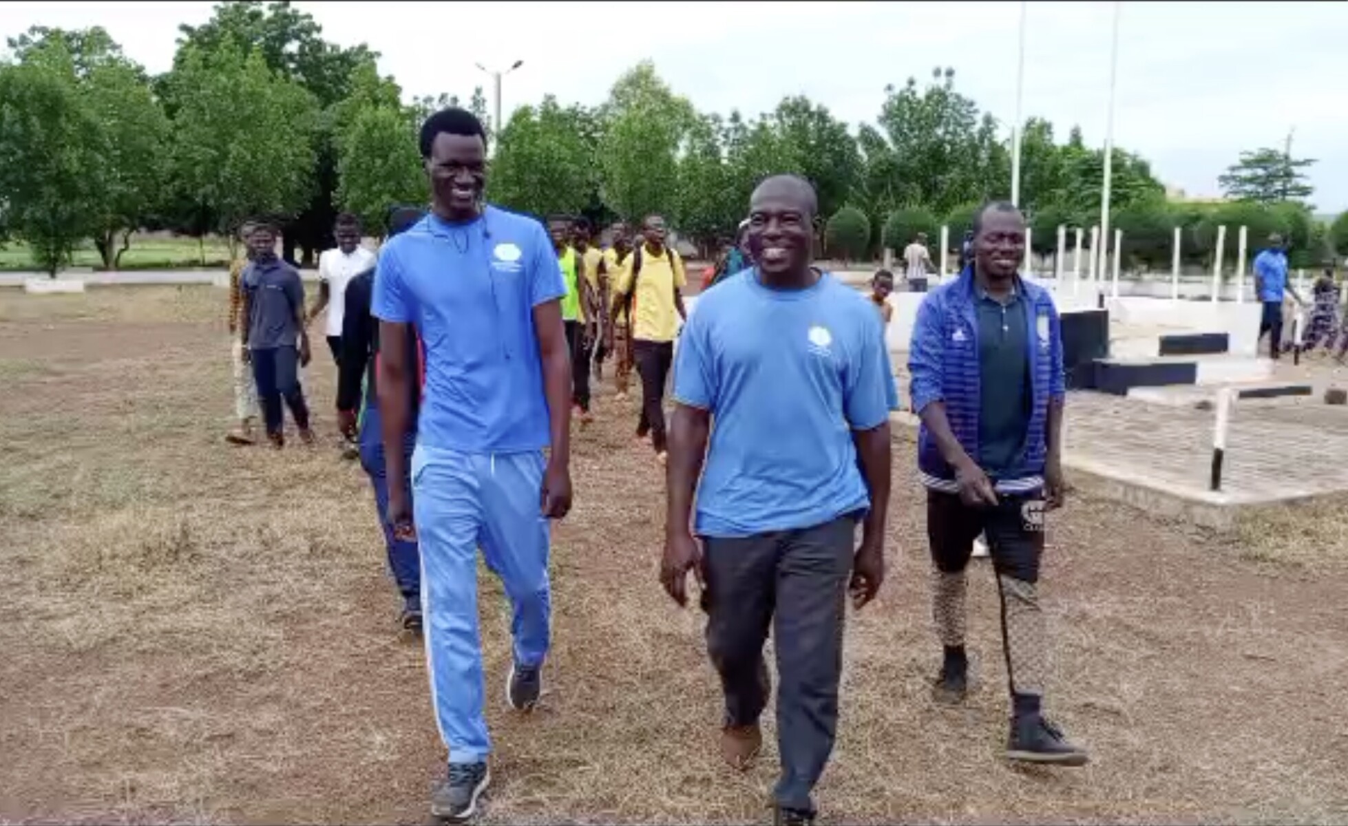 Three men lead a group of people walking outside. They wear blue shirts and they smile.