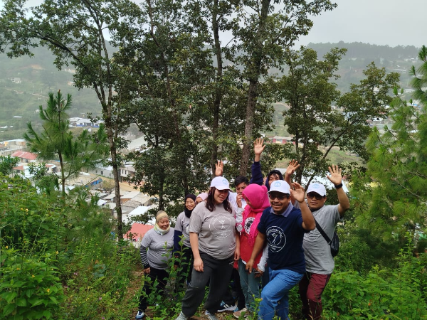 People wave while walking on a hill with bushes and trees. Houses are visible down the hill in the background.