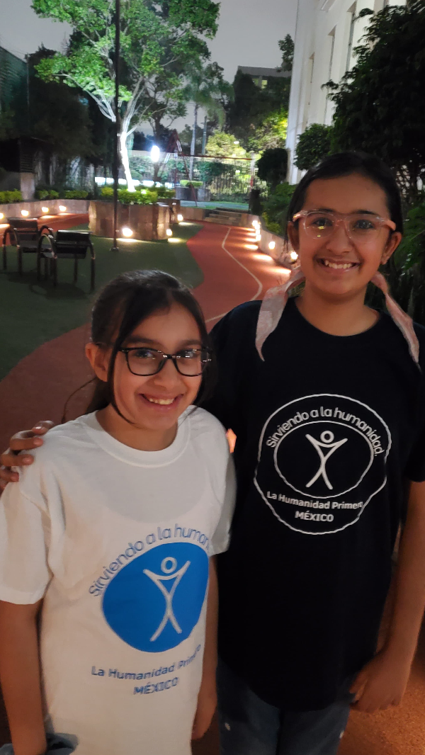 Two girls stand side by side on a sidewalk at night. One wears a blue shirt and the other a white shirt both with humanity first logos.