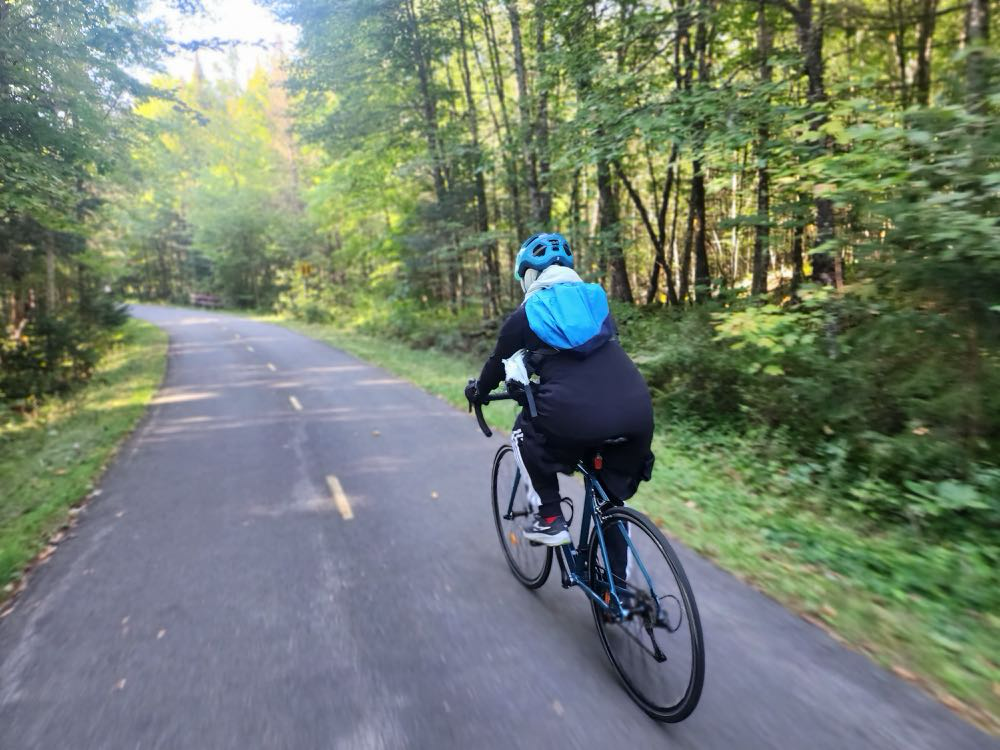 A woman on a bike wearing a bright blue helmet and backpack rides on a paved bike path through the woods