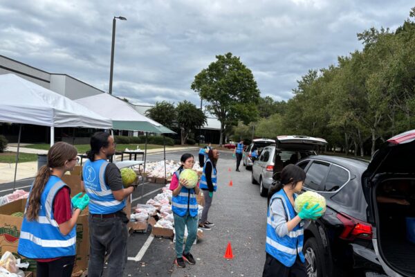 People in blue vests put items like watermelon in the open trunks of cars that wait in line for food
