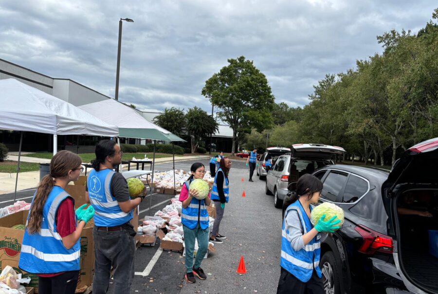 People in blue vests put items like watermelon in the open trunks of cars that wait in line for food