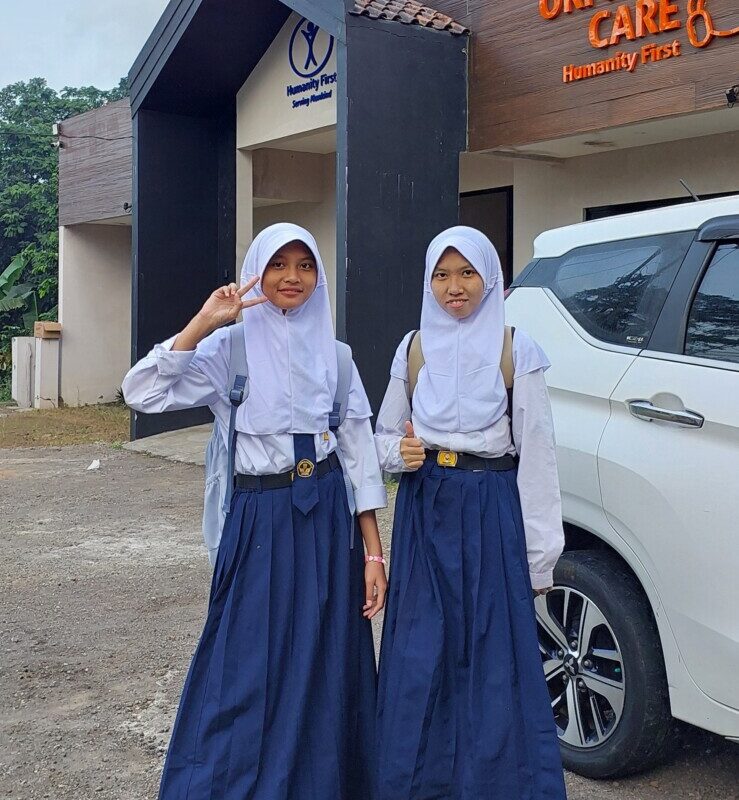 Two girls stand in front of the Humanity First orphanage. They wear matching school uniforms of long blue skirts white tops and white hijab. One makes a peace sign and the other gives a thumbs up.