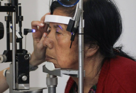 Elderly woman sits with her face in an eye measurement machine above the text Gift of Sight