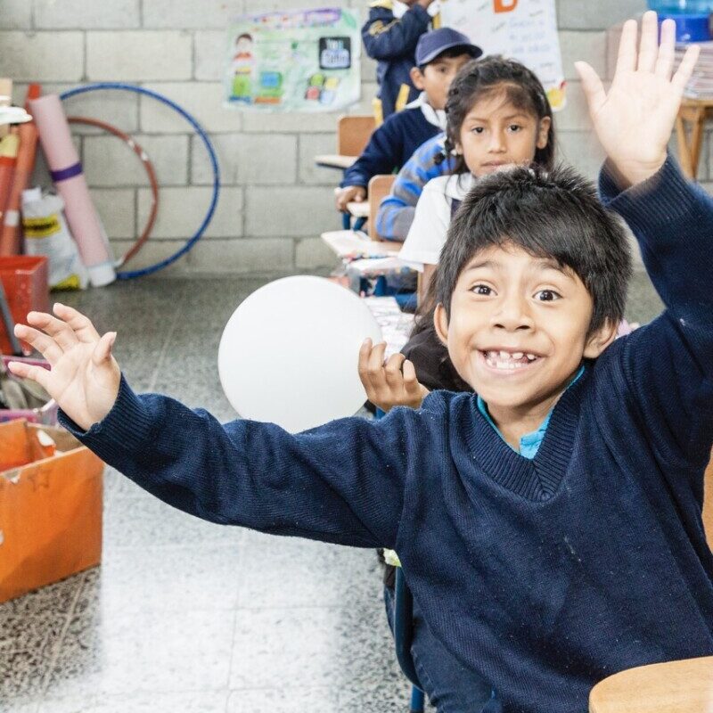 A young boy sitting at the front of a row of school desks gives a huge smile while waving his arms in the air.