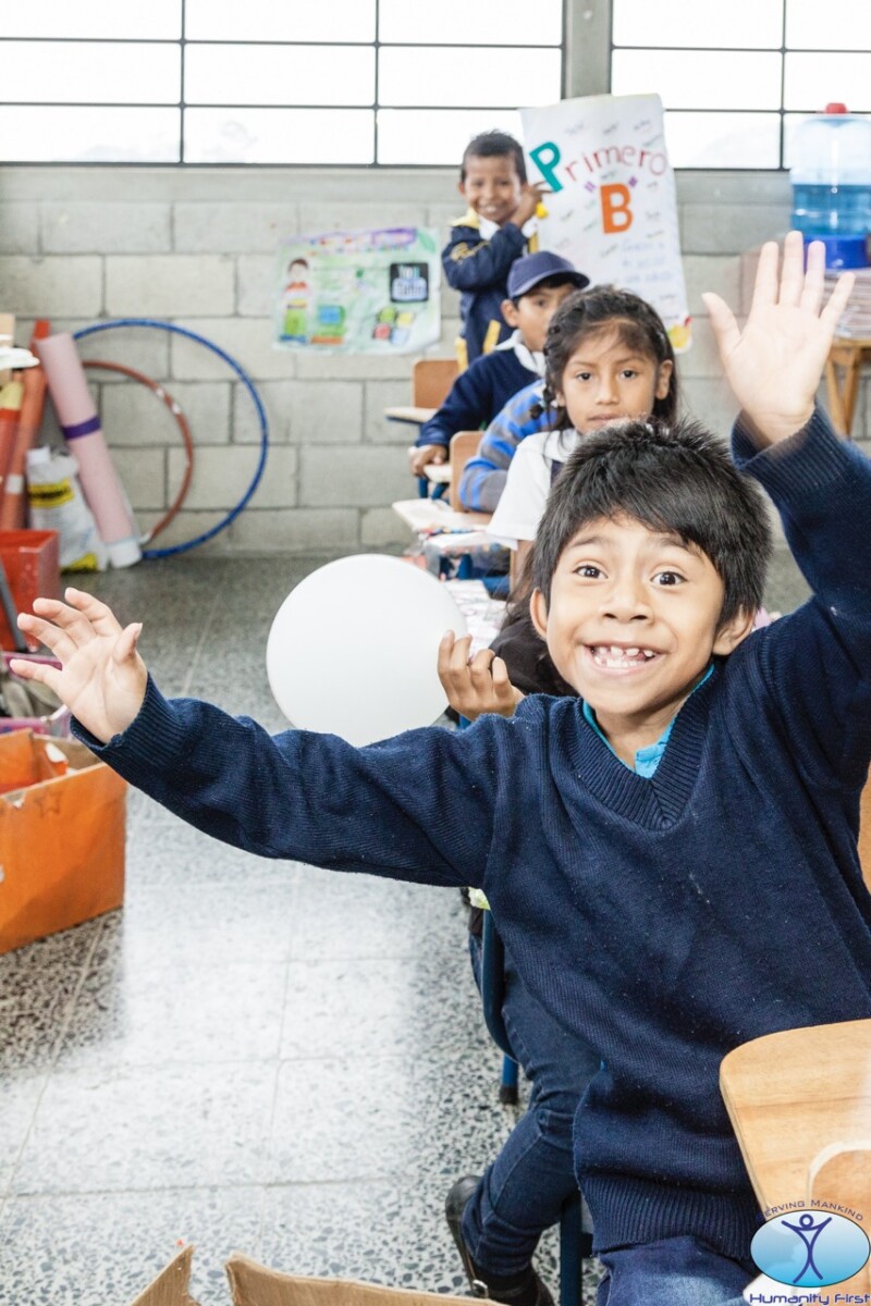 A young boy sitting at the front of a row of school desks gives a huge smile while waving his arms in the air.