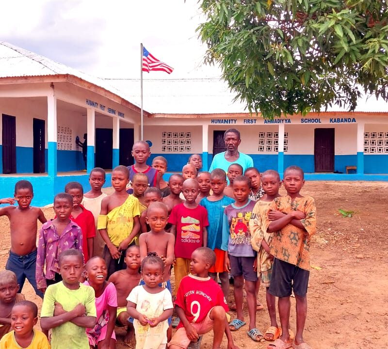 A group of young students wearing t shirts and shorts standing with a teacher in front of a one story school building painted blue and white.