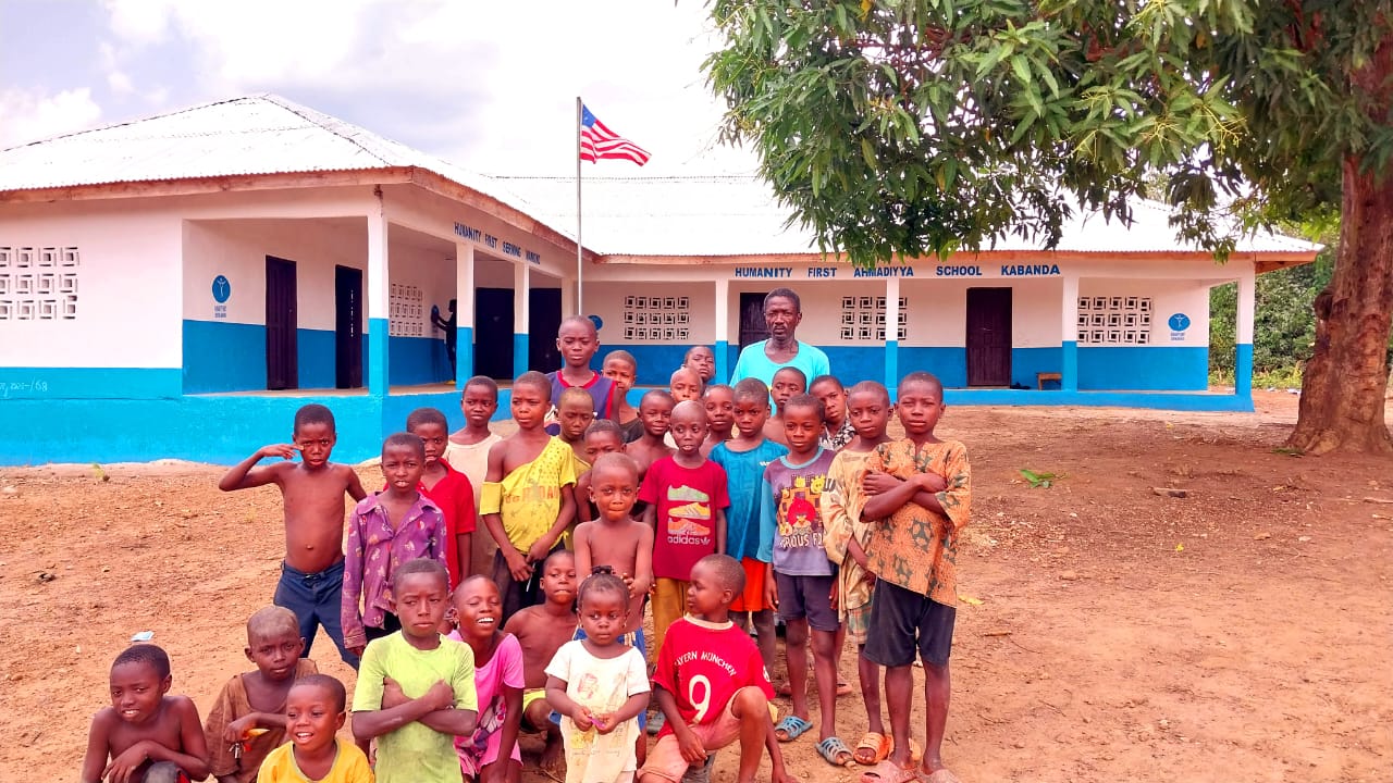 A group of young students wearing t shirts and shorts standing with a teacher in front of a one story school building painted blue and white.