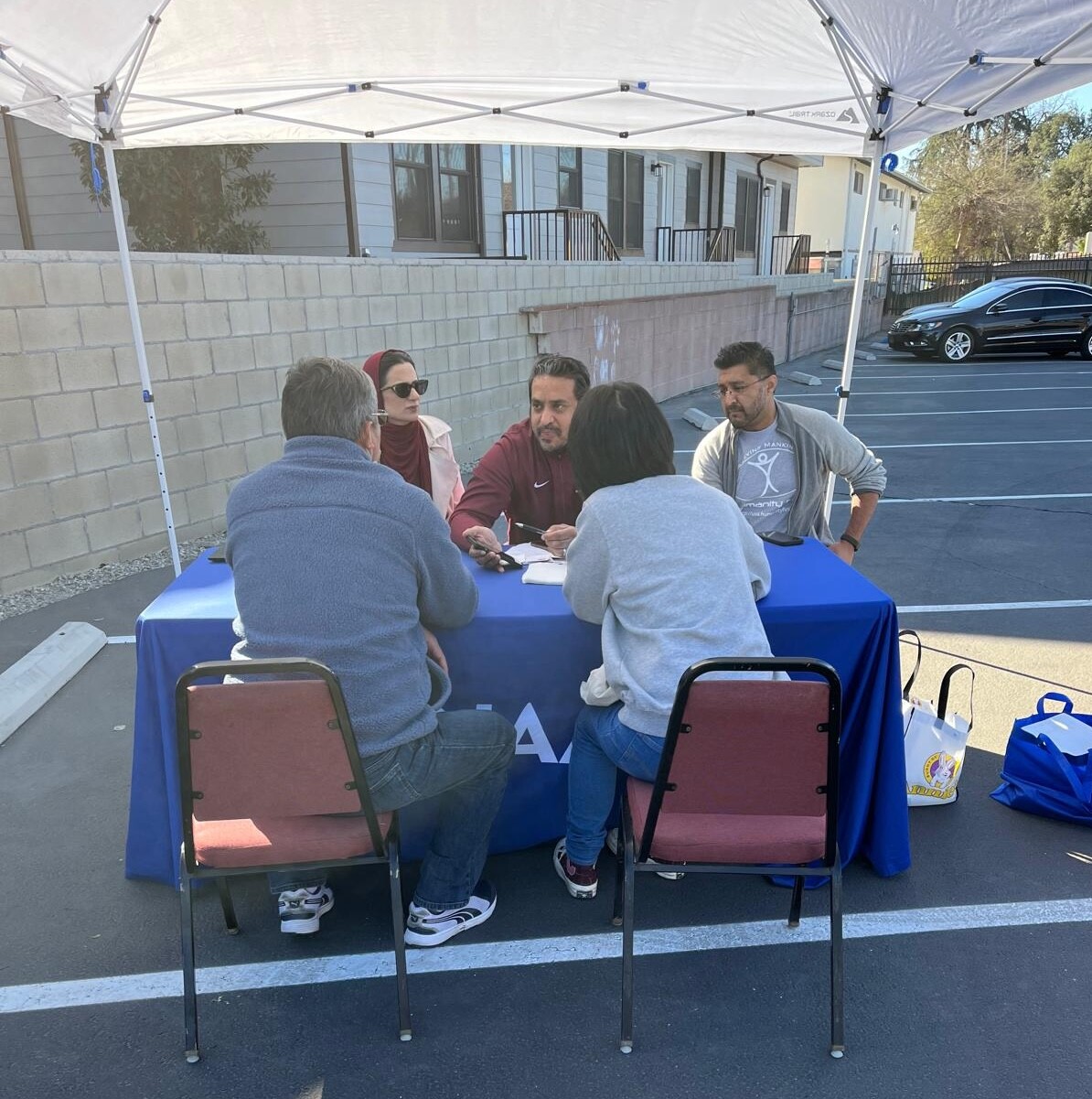 five people sit opposite each other across a table under an open air tent in a parking lot