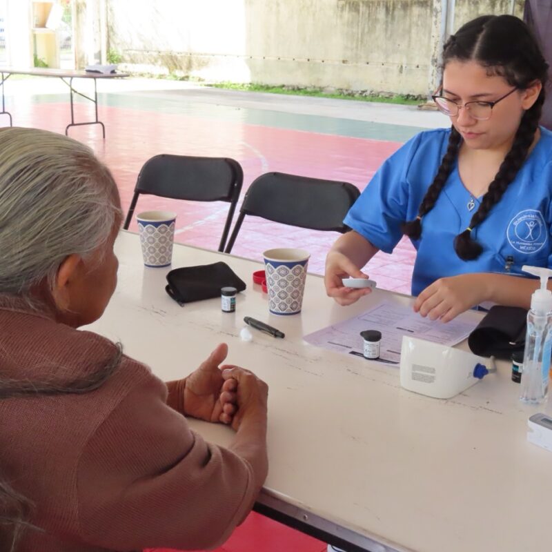 A woman wearing blue medical scrubs sits opposite an elderly woman and looks at a blood glucose reader.