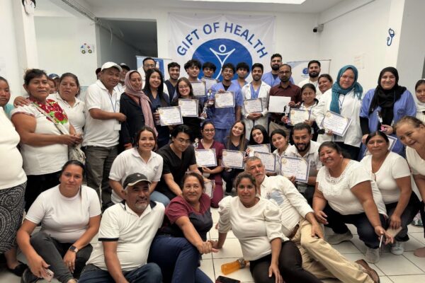 A couple dozen people pose in front of a sign that says Gift of Health. A set of post-secondary students in the middle hold certificates.