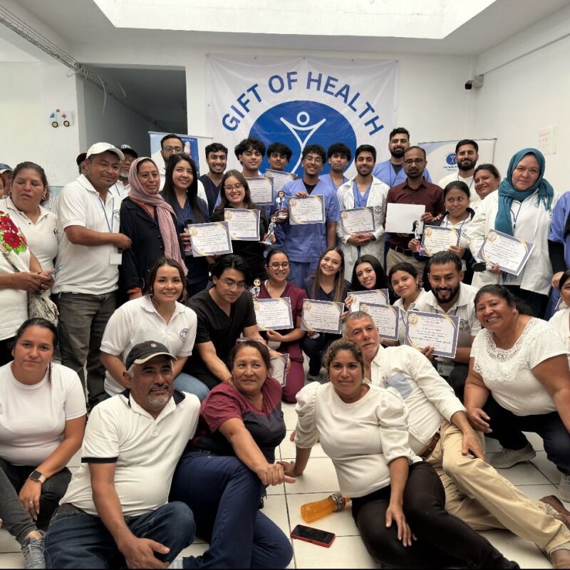 A couple dozen people pose in front of a sign that says Gift of Health. A set of post-secondary students in the middle hold certificates.