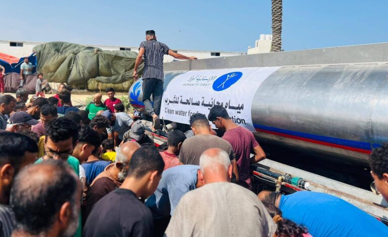 people crowd around a water tanker that bears the humanity first logo. one man walks on the side of the tanker overlooking the people getting water.