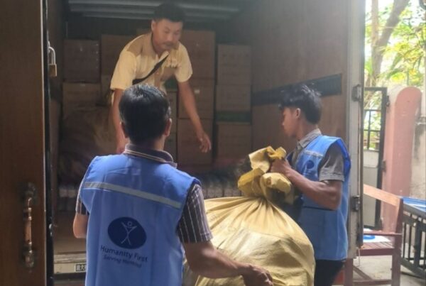 Three men wearing blue vests load a very large burlap bag into the open back of a truck that has boxes already piled in the back.