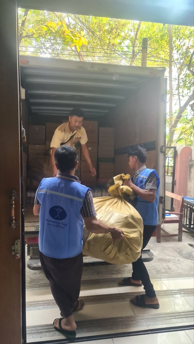 Three men wearing blue vests load a very large burlap bag into the open back of a truck that has boxes already piled in the back.