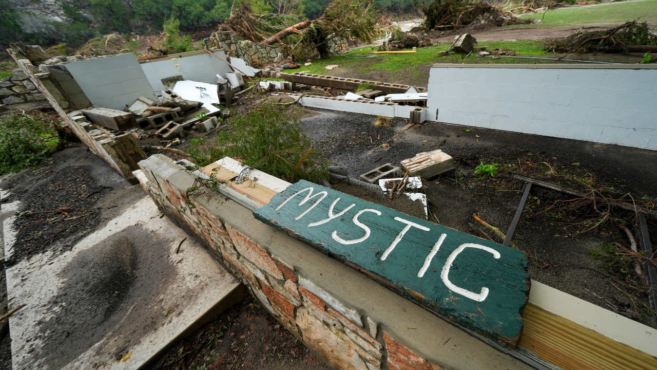 An image of fallen trees and a building washed away. A short wall remains with a sign resting on top of it reading Mystic.
