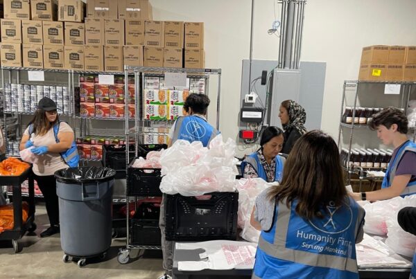 People wearing blue humanity first vests pack food at different stations in the HF Duluth Food Pantry warehouse that is packed with food items on shelves and stacked boxes.