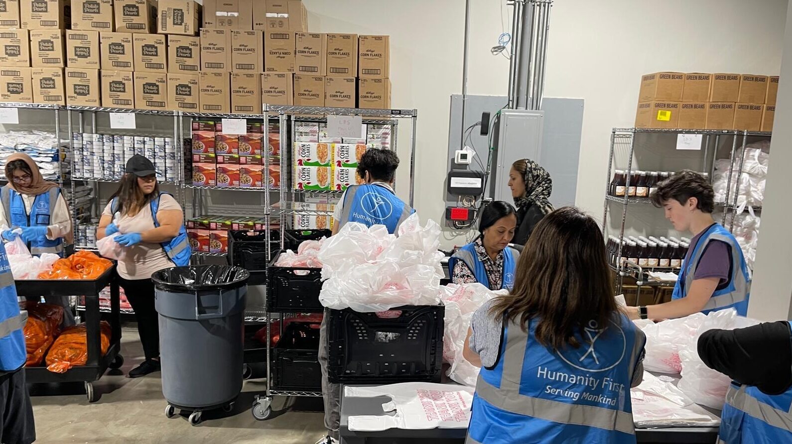 People wearing blue humanity first vests pack food at different stations in the HF Duluth Food Pantry warehouse that is packed with food items on shelves and stacked boxes.