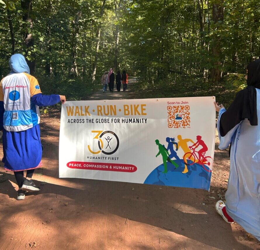 Two ladies, one on each side, hold a Humanity First 30th Anniversary Walk Run Bike event banner. They are walking on a path in the woods and others in their group walk ahead of them.