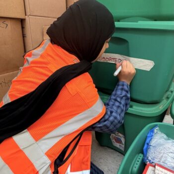 A woman wearing an high visibility vest labels large plastic packing containers full of relief supplies.