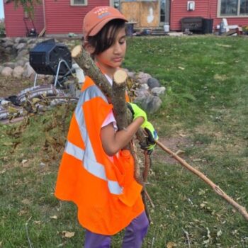 A child wearing a high visibility vest picks up small tree branches from a yard.