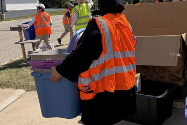 A woman wearing a high visibility vest carries a plastic tub of relief supplies out to the street.