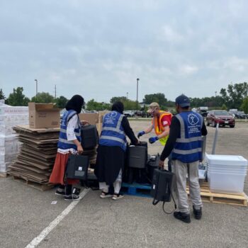 Three people wearing blue Humanity First vests unload supplies from pallets in a parking lot.