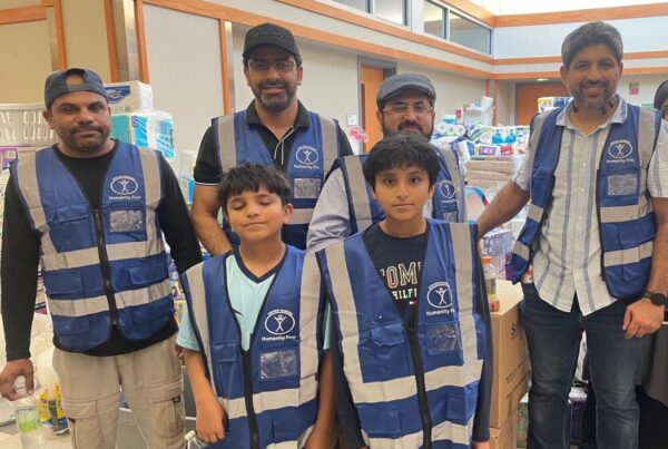 A group of men and boys wearing blue Humanity First vests stand with boxes of packed hygiene and home supplies for disaster relief.